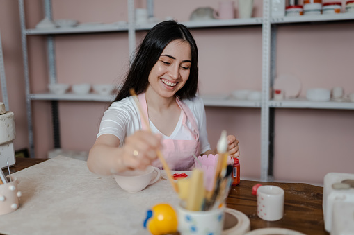 Beautiful young woman is painting a ceramic mug in workshop