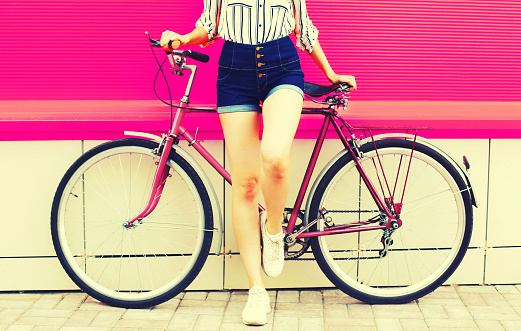 Summer image of legs of beautiful young woman in shorts posing with bicycle in the city on pink background
