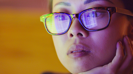 Close-up image of an Asian woman watching a computer screen of numbers which are reflected in her glasses.