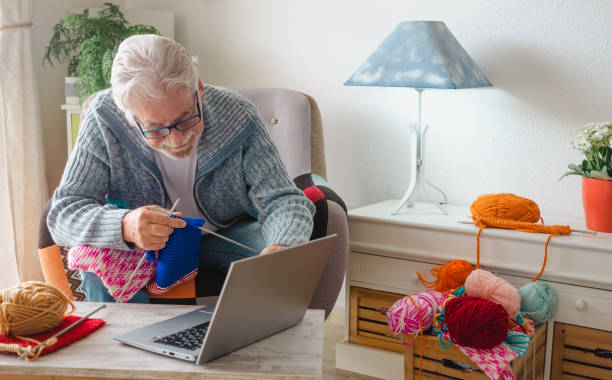 istock Smiling senior man enjoying knitting at home following online knit tutorial to relax and enjoy leisure activity sitting on armchair - male people knitting with computer class help 1404392654