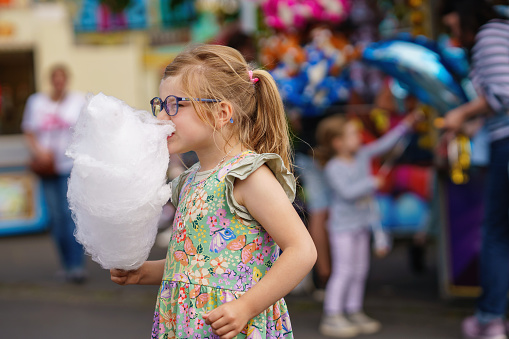 Adorable little girl eating cotton candy in amusement park. Happy preschool child with glasses and big candy-floss, outdoors on summer sunny day