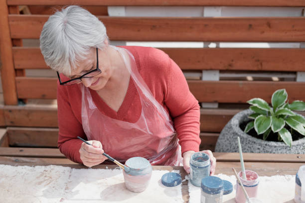 眼鏡をかけた年配の白髪の女性は、手作りの粘土の花瓶をブラシで塗っています。 - hobbyist ストックフォトと画像