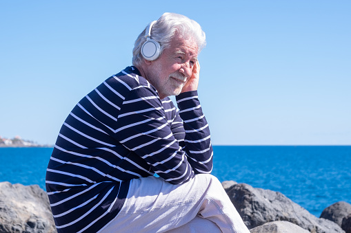 Caucasian elderly man with headphones sitting on the cliffs enjoying sea and freedom in vacation