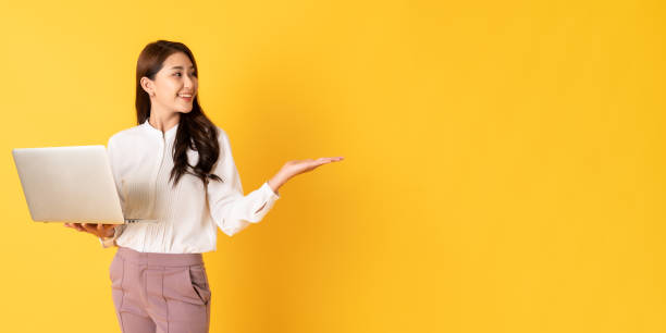 Young attractive woman in smart casual cloth using laptop on yellow background presenting object on copy space