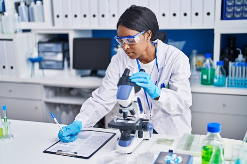 Young african american woman scientist using microscope write on document at laboratory