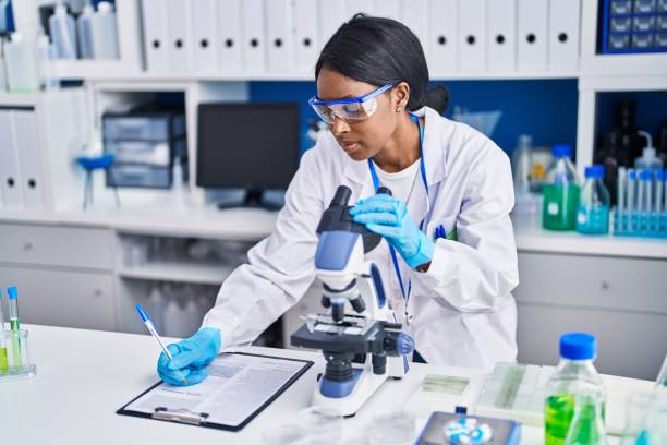 Young african american woman scientist using microscope write on document at laboratory stock photo