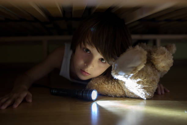 istock Little child, hiding under the bed, hugging teddy bear and holding flashlight, 1404417398