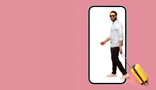 Great mobile app for tourism and travel. Cheerful carefree Indian man in sunglasses walking with luggage into huge smart phone isolated on pink, advertising of booking platform, copy space, mock up
