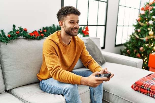 istock Young arab man playing video game sitting on the sofa by christmas tree at home. 1404422866