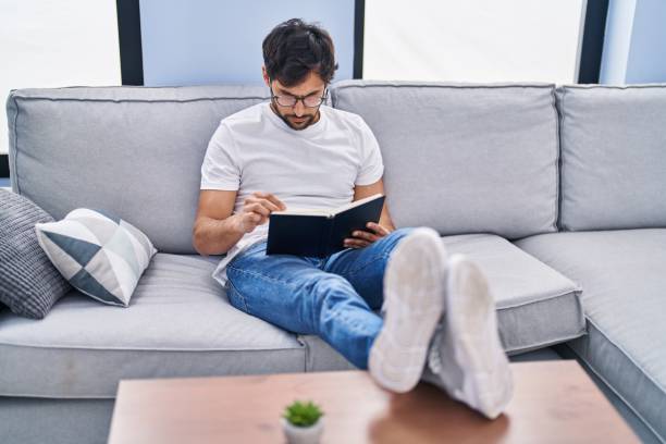 istock Young hispanic man reading book sitting on sofa at home 1404423275