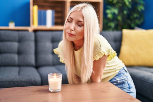 jeune femme blonde sentant l’odeur d’une bougie assise sur un canapé à la maison - femme sentir bougie photos et images de collection