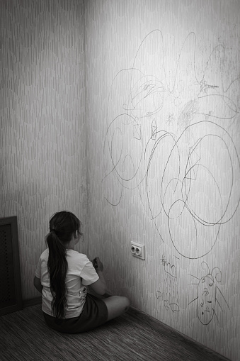 Back view of little naughty girl with dark hair wearing T-shirt, holding green marker in hand, sitting on floor, drawing picture of bug on old yellow wallpaper. Misbehavior, redecoration. Vertical.