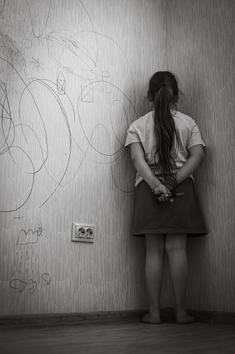 Black and white portrait of little bad girl with dark hair holding marker behind back, standing punished in corner for spoilt wallpaper, facing wall. Misbehavior, punishment, home violence. Vertical.