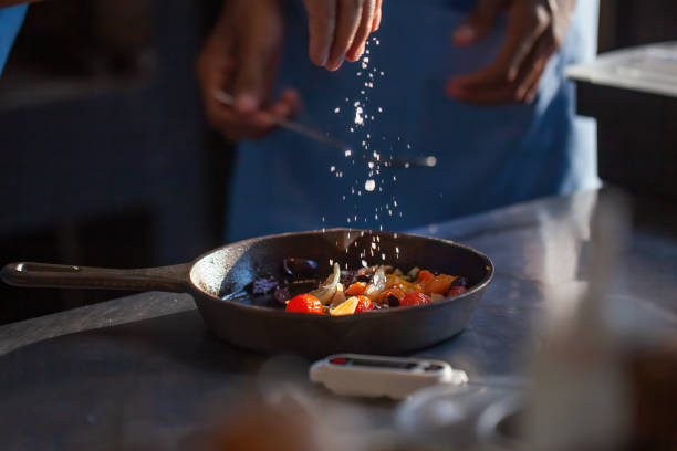 istock Chef cook preparing vegetables in luxury restaurant or hotel kitchen. Man hands add salt in roasted vegetables in pan. Professional cook in uniform add some species to dish. 1404430927