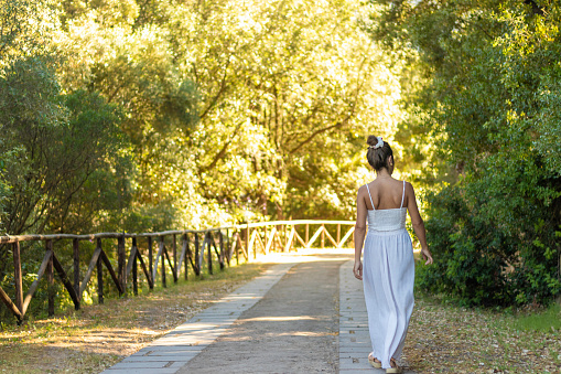 Unrecognizable backward young brunette woman looking to the side in white summer outfit walking along nature in an empty path