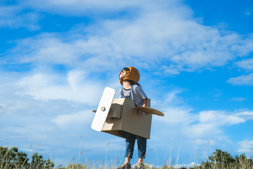 Cute dreamer little girl playing with cardboard planes in the meadow on a sunny day. Happy kid playing with cardboard plane against blue summer sky background. Childhood dream imagination concept.