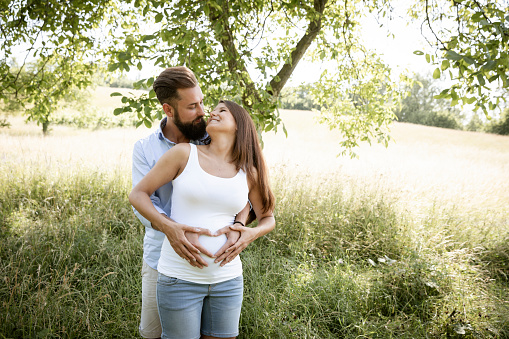 pretty young pregnant woman with white shirt stands with her boyfriend with beard and blue shirt in high flower meadow and cuddle