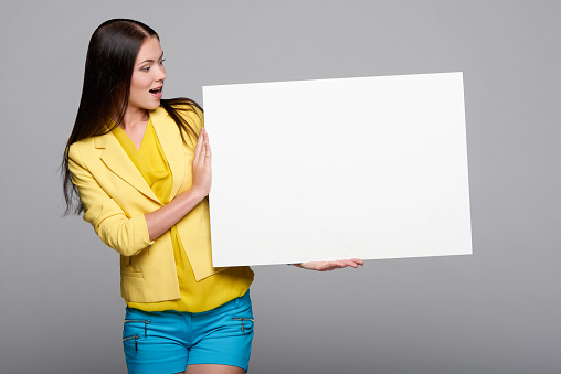 Surprised girl in yellow and blue, holding empty whiteboard, looking at blank copy space at board