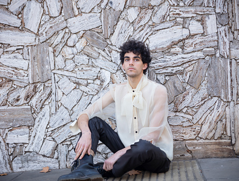 Portrait of a young LGBTQ+ man with stilysh haircut and clothes in front of wall in street