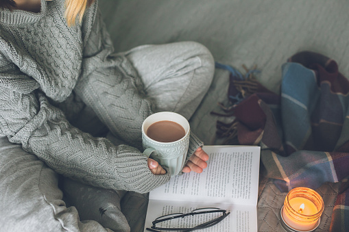 Winter still life concept. Young girl in knitted warm clothes holding cacao mug, reading book. Season holiday, hygge concept