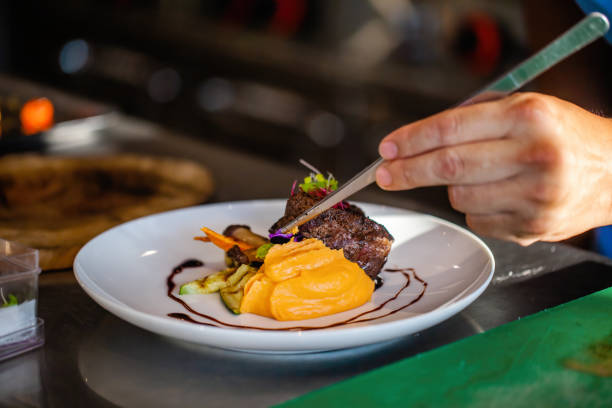 istock Close up of chef hands in hotel or restaurant kitchen decorating dish. Preparing beef steak with mashed potato, adding vegetables, decorate dish with special tweezers. Chef hands cooking meat steak. 1404453123