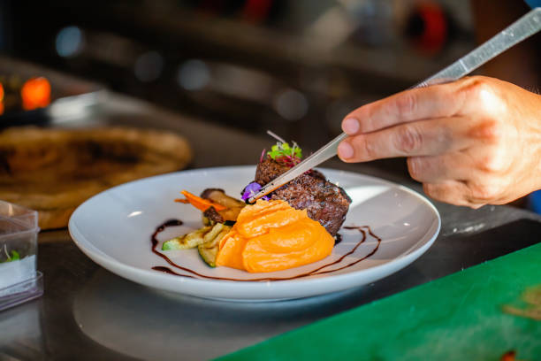 istock Close up of chef hands in hotel or restaurant kitchen decorating dish. Preparing beef steak with mashed potato, adding vegetables, decorate dish with special tweezers. Chef hands cooking meat steak. 1404459598