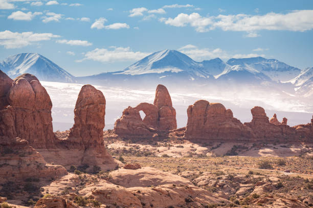 A lonely car driving through the Arches National Park. Behind the dry desert there are snow capped mountains. Seen at day in the summer.