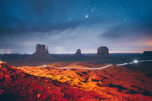 Night sky with stars showing on a dramatic sky above the towering sand stone buttes at Monument Valley. Located on the Arizona and Utah border of the southwest USA. Glowing lights showing from a car passing by on the gravel road.