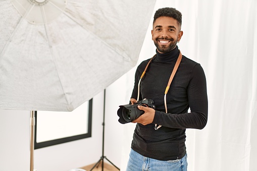 Handsome hispanic man working as professional photographer at photography studio. Standing holding camera smiling confident.