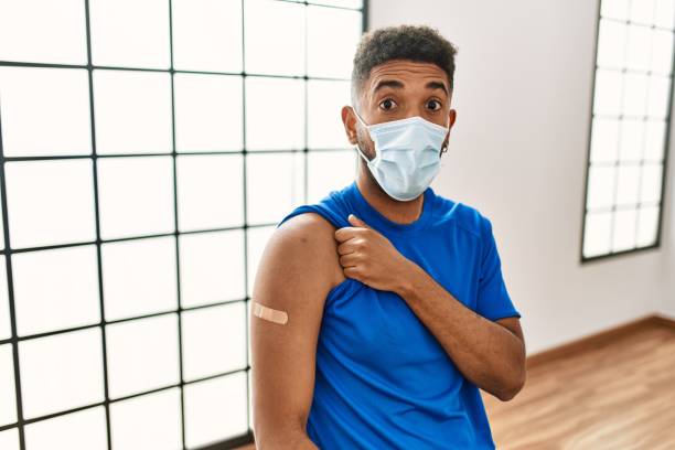 istock Young hispanic man with beard wearing safety mask getting vaccine scared and amazed with open mouth for surprise, disbelief face 1404468318