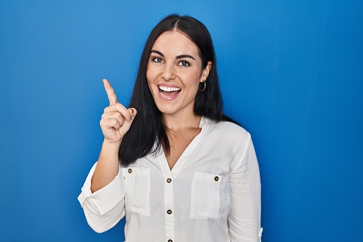 Young hispanic woman standing over blue background pointing finger up with successful idea. exited and happy. number one.