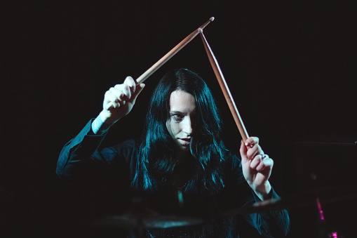 A female metal drummer with long hair holding up drumsticks behind the drumkit against a dark background