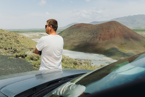 Young man explorer contemplating the road trip by a car through the scenic volcanic landscape with a colorful crater and the Meke Lake