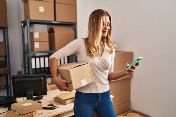 istock Young blonde woman ecommerce business worker using smartphone holding package at office 1404473584