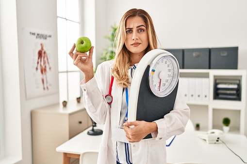 Young blonde doctor woman holding weighing machine and green apple skeptic and nervous, frowning upset because of problem. negative person.