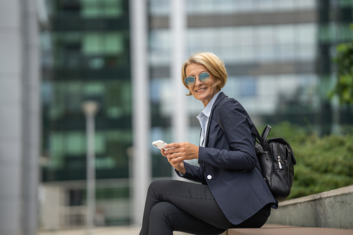 Beautiful mature business woman texting message on mobile phone outside business center on sunny day
