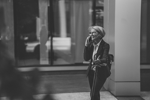 Black and white shot of mature business woman with push scooter standing in front of office building and talking on mobile phone