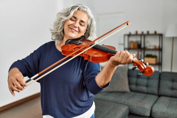 Middle age grey-haired artist woman playing violin standing at home. stock photo