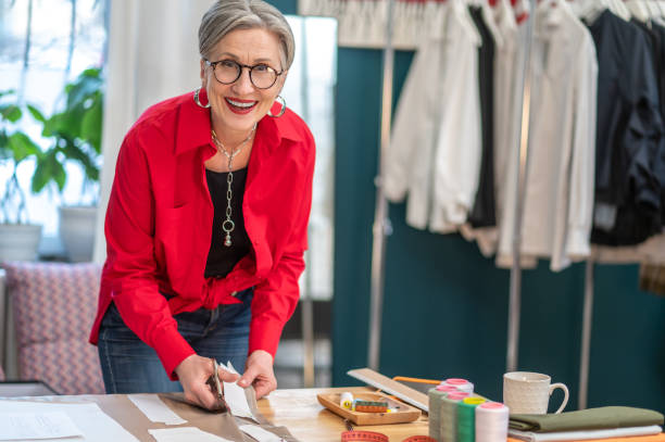 Happy woman looking at camera making pattern Lucky day. Happy middle aged woman in glasses looking confidently at camera standing near table cutting out pattern in atelier middle aged woman fashion stock pictures, royalty-free photos & images