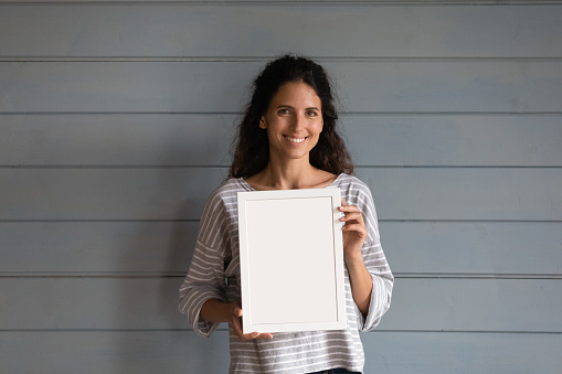 Happy pretty Hispanic 30s woman with curly hair holding blank empty white picture frame with copy space for text, standing at grey wall background, looking at camera. Head shot portrait