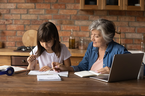 Senior grandmother helping schoolkid granddaughter to do homework. Grandma, nanny, teacher watching explaining writing exercise to girl, pointing at copybook, speaking at desk with laptop