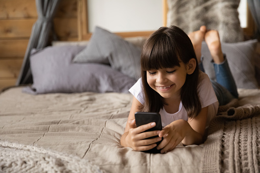 Smiling pretty kid using smartphone at home, relaxing on bed in bedroom, enjoying communication, friendly talk on video call. Happy gen Z girl using online app on mobile phone, making telephone call