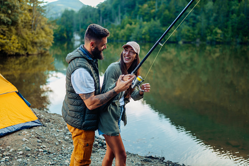 Male trying to teach his girlfriend how to fish with a fishing rod by a lake