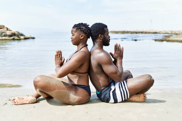 Young african american tourist couple wearing swimwear meditating doing yoga exercise at the beach. stock photo