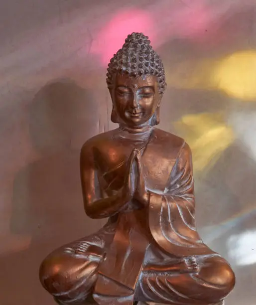 Golden buddha statuette with hands folded like the Indian Namaste greeting (Anjali mudra). He sits in pink and yellow light reflections coming from objects in the window.