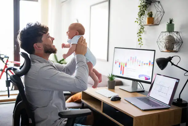 Multi-tasking father taking care of baby son while working at home office.