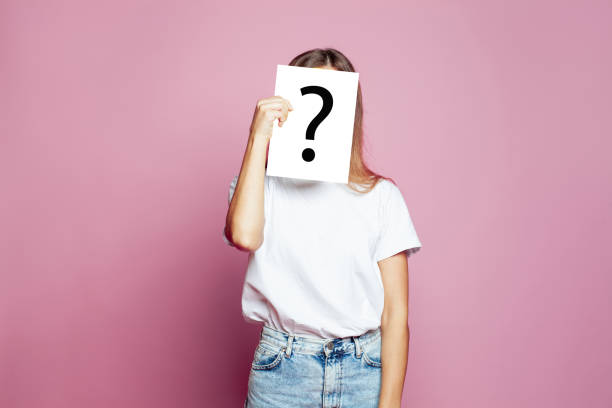 istock Pretty woman covering face using a white paper sheet with drawn question mark, like a mask, for hiding her identity on pink background 1404498078