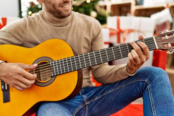 istock Young hispanic man playing guitar sitting by christmas tree at home 1404498980