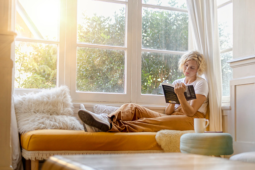 A young mother unwinds after a long day by reading her favourite book. She is seated crossed legged in a reading nook in a bright and airy lounge room. The sun is setting and shining through the large windows.