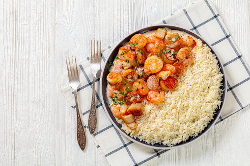 shrimps and scallops in spicy coconut cream sauce with long grain rice on plate on white wooden table with forks,  horizontal view from above, flat lay, free space